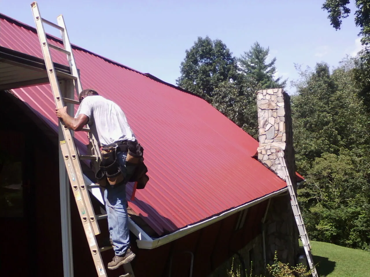 Roofer on a ladder beside a red metal roof and stone chimney — Soffit & Fascia Repair by Roofing Brothers in Plainwell